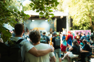 Zwei Besucher stehen umarmt im Vordergrund, während auf der Bühne des Elbenwald Festivals eine Band spielt. Im Hintergrund sind zahlreiche Festivalbesucher in bunten Kostümen zu sehen, die die lebhafte Atmosphäre genießen.