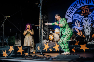 Opening mit Olaf Unzel und SaKiDe Zwei Performer auf der Bühne des Elbenwald Festivals, gekleidet in auffälligen Kostümen. Die Frau mit lila Haaren hält ein Mikrofon, während der Mann in einem grünen Anzug mit Fragezeichen animiert auftritt. Im Hintergrund sind Schlagzeug und Festivalbanner zu sehen.