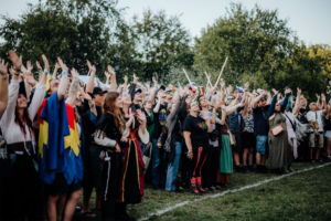 Eine begeisterte Menge von Festivalbesuchern hebt die Hände in die Luft, umgeben von einer bunten Atmosphäre beim Elbenwald Festival. Die Teilnehmer sind in kreativen Kostümen gekleidet und genießen die lebendige Stimmung des Events.