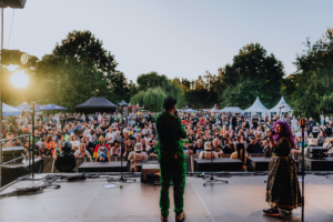 Opening mit Olaf Unzel und SaKiDe Bühne beim Elbenwald Festival mit einem Publikum von begeisterten Fans. Ein Künstler in grünem Anzug steht auf der Bühne, während eine Sängerin mit lila Haaren das Publikum anfeuert. Im Hintergrund sind Zelte und Bäume sichtbar, die die Festivalatmosphäre unterstreichen.