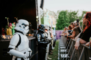 Stormtrooper-Kostüme stehen an der Bühnenfront beim Elbenwald Festival. Festivalbesucher beobachten gespannt die Darbietungen auf der Bühne, während die Atmosphäre von Musik und Nerdkultur geprägt ist.