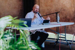 Parakryptozoologie mit Christian von Aster Ein Redner in einem weißen Kittel präsentiert auf dem Elbenwald Festival an einem Tisch mit Laptop und Mikrofon. Im Hintergrund sind Pflanzen sichtbar, während er gestikuliert und das Publikum anspricht.