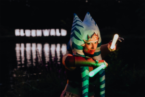 Cosplayerin in fantasievoller Kleidung mit leuchtenden Stäben auf dem Elbenwald Festival, im Hintergrund das beleuchtete Festival-Logo. Die Szenerie spiegelt eine kreative Mischung aus Musik und Nerdkultur wider, umgeben von einem malerischen Gewässer.