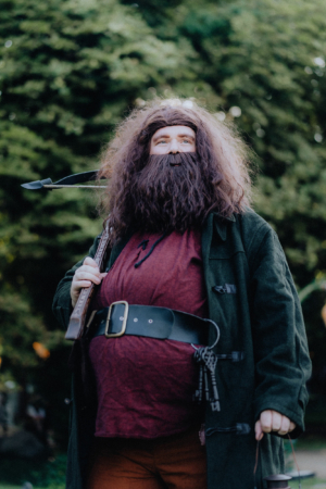 Ein kostümierter Besucher des Elbenwald Festivals posiert mit einem Gewehr und einer Laterne. Der Mann trägt ein rotes Shirt, eine grüne Jacke und hat einen langen, lockigen Bart. Im Hintergrund sind verschwommene Bäume und Festivalbesucher zu sehen.