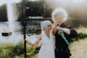 Cosplay-Paar auf dem Elbenwald Festival, umgeben von einer malerischen Kulisse mit einem Teich und Sprühfontäne. Die beiden Charaktere tragen aufwendige Kostüme, die kreative Designs und fantasievolle Accessoires präsentieren, während der Schriftzug "Elbenwald" im Hintergrund sichtbar ist.