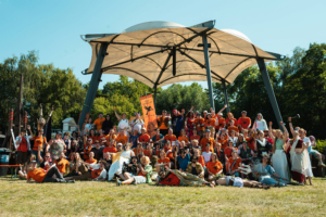 Percy Jackson Talk Gruppenselfie von Teilnehmern des Elbenwald Festivals, die in bunten Kostümen und orangefarbenen T-Shirts posieren. Die fröhlichen Festivalbesucher stehen vor einem modernen Pavillon und genießen die kreative Atmosphäre des Events.