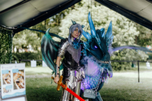 Ryu's Drachenhöhle Ein Cosplayer in einer beeindruckenden Rüstung mit Drachenflügeln steht neben einem leuchtenden Drachen. Die Szene zeigt die kreative Atmosphäre des Elbenwald Festivals, das Musik und Nerdkultur vereint.