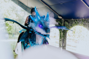 Ein kostümierter Teilnehmer reitet auf einem beeindruckenden blauen Drachen mit leuchtenden Augen und Nebel-Effekten, umgeben von einer festlichen Zeltatmosphäre beim Elbenwald Festival.