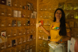 Elbenwald Festival Post Eine lächelnde Frau in einem gelben T-Shirt steht in einem Stand auf dem Elbenwald Festival, umgeben von Postkarten in transparenten Fächern. Sie hält eine Postkarte in der Hand und präsentiert stolz die Auswahl an Karten.