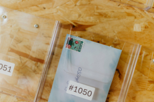 Elbenwald Festival Post Ein Briefumschlag mit einer bunten Briefmarke, der in einem transparenten Behälter auf dem Elbenwald Festival platziert ist. Der Umschlag ist mit der Nummer #1050 gekennzeichnet und vermittelt das kreative, nerdige Flair des Festivals.