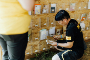 Elbenwald Festival Post Ein Teilnehmer des Elbenwald Festivals schaut sich handgeschriebene Briefe in einem dafür vorgesehenen Bereich an, während eine andere Person im gelben T-Shirt im Hintergrund zu sehen ist. Die Wand ist mit transparenten Taschen für die Briefe ausgestattet.