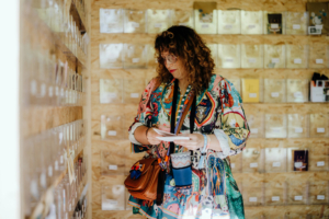 Elbenwald Festival Post Eine Frau mit lockigem Haar und buntem Kleid steht vor einer Wand mit transparenten Fächern, die verschiedene Karten und Merchandise-Artikel zeigen. Sie hält Zettel in der Hand und betrachtet die Auslage aufmerksam auf dem Elbenwald Festival.