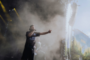 Punk Rock Factory Ein Musiker steht auf der Bühne des Elbenwald Festivals, umgeben von Nebel und bunten Lichtern. Im Hintergrund sind weitere Bandmitglieder und ein großes Festivalbanner sichtbar, das die kreative und lebendige Atmosphäre des Events unterstreicht.