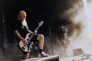 Punk Rock Factory Zwei Musiker auf der Bühne des Elbenwald Festivals, umgeben von Nebel. Der Bassist mit Bart spielt ein Instrument, während der Sänger leidenschaftlich ins Mikrofon singt. Die energiegeladene Atmosphäre und das Publikum im Hintergrund schaffen ein unvergessliches Erlebnis.
