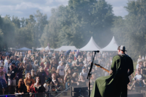 Punk Rock Factory Ein Musiker auf der Bühne des Elbenwald Festivals, umgeben von einer begeisterten Menschenmenge. Die Atmosphäre ist lebhaft und festlich, mit Zelten im Hintergrund und einer Vielzahl von Kostümen im Publikum. Ideal für Fans von Musik und Popkultur.