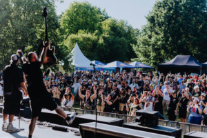 Punk Rock Factory Bühne beim Elbenwald Festival mit performenden Musikern und begeistertem Publikum. Die Zuschauer feiern inmitten von Bäumen und Festzelten, während die Atmosphäre von Musik und Freude erfüllt ist. Ein perfekter Moment der Gemeinschaft und des Spaßes.