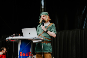 Quiz mit Olaf Unzel Ein Mann im Kostüm eines berühmten Videospielcharakters steht an einem Rednerpult mit einem Laptop. Im Hintergrund sind verschwommene Festivalbesucher zu sehen. Die Atmosphäre ist lebhaft und kreativ, typisch für das Elbenwald Festival.