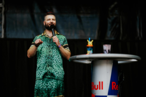 Quiz Ein Moderator in einem lebhaften grünen Outfit steht auf der Bühne des Elbenwald Festivals und spricht ins Mikrofon. Auf dem Tisch neben ihm steht eine bunte Pikachu-Trinkflasche, die die nerdige Atmosphäre des Events unterstreicht.