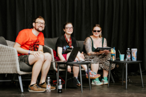 Quiz Drei Festivalbesucher sitzen entspannt auf einer Couch während des Elbenwald Festivals. Sie lächeln, während sie an einem Tisch mit Getränken und einem Laptop sitzen. Die Atmosphäre ist freundlich und lässig, ideal für eine Mischung aus Musik und Nerd-Kultur.