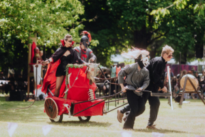 Ompah Kunstreitshow Künstlerische Darbietung beim Elbenwald Festival: Eine Gruppe von Darstellern in fantasievollen Kostümen zieht eine rote Kutsche, während sie eine lebhafte Szene im Freien aufführen. Die Veranstaltung verbindet Musik und Nerd-Kultur in einer kreativen Atmosphäre.