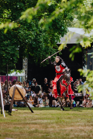 Ompah Kunstreitshow Ein Ritter in roter Rüstung reitet auf einem Pferd und schwingt einen Speer während einer Vorführung auf dem Elbenwald Festival. Im Hintergrund sind zahlreiche Zuschauer zu sehen, die gespannt die Darbietung verfolgen, umgeben von grünen Bäumen.