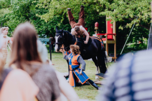Ompah Kunstreitshow Darstellung einer Aufführung auf dem Elbenwald Festival, bei der Künstler auf einem Pferd akrobatische Kunststücke vorführen. Im Vordergrund sind Zuschauer zu sehen, die gebannt zuschauen. Die Atmosphäre ist lebhaft und kreativ, geprägt von Fantasie und Gemeinschaft.