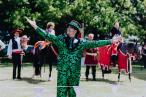 Ompah Kunstreitshow Ein farbenfroher Performer in einem grünen Anzug präsentiert sich mit ausgebreiteten Armen auf dem Elbenwald Festival. Im Hintergrund stehen weitere Darsteller in historischen Kostümen und auf Pferden, die die festliche Atmosphäre unterstreichen.