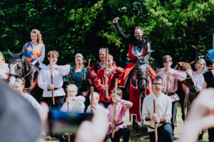 Ompah Kunstreitshow Eine Gruppe von Teilnehmern in historischen Kostümen posiert beim Elbenwald Festival, darunter ein Reiter in Rüstung auf einem Pferd und Darsteller mit Stöcken. Die Szenerie ist von grüner Natur umgeben und vermittelt ein festliches, lebhaftes Ambiente.