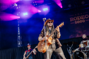 Rodeo 5000 Ein Musiker mit Dreadlocks und Sonnenbrille spielt Gitarre auf der Bühne des Elbenwald Festivals. Im Hintergrund sind Bandkollegen und farbige Lichter zu sehen, die die energiegeladene Atmosphäre des Festivals unterstreichen.