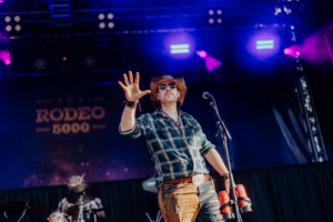 Rodeo 5000 Ein Musiker in Cowboy-Hut und Sonnenbrille auf der Bühne des Elbenwald Festivals, während er mit einer Hand das Publikum anspricht. Im Hintergrund sind bunte Lichter und ein Schlagzeuger sichtbar. Die Atmosphäre ist lebhaft und energiegeladen.