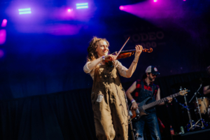 Rodeo 5000 Eine Musikerin spielt Geige auf der Bühne des Elbenwald Festivals, umgeben von bunten Lichtern. Im Hintergrund ist ein weiterer Musiker mit Bassgitarre zu sehen. Die Atmosphäre ist lebhaft und festlich, ideal für Fans von Musik und Nerdkultur.