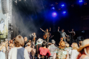Rodeo 5000 Auf der Bühne des Elbenwald Festivals performt eine Band vor einer begeisterten Menge. Musiker mit Gitarre und Violine sorgen für eine lebendige Atmosphäre, während das Publikum in fantasievollen Kostümen feiert. Bunte Lichter und Nebel schaffen eine festliche Stimmung.