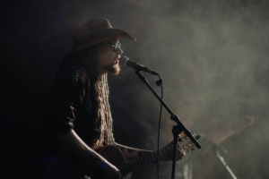 Rodeo 5000 Ein Musiker mit langen Dreadlocks und Cowboyhut spielt Gitarre und singt vor einem Mikrofon auf der Bühne des Elbenwald Festivals, umgeben von Nebel. Die Atmosphäre ist lebhaft und kreativ, perfekt für Fans von Musik und Nerd-Kultur.