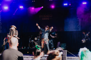 Rodeo 5000 Auftritt der Band Rodeo 5000 auf dem Elbenwald Festival, umgeben von begeisterten Fans. Die Bühne ist mit bunten Lichtern beleuchtet, während die Musiker mit Instrumenten und Gesang eine energiegeladene Atmosphäre schaffen.
