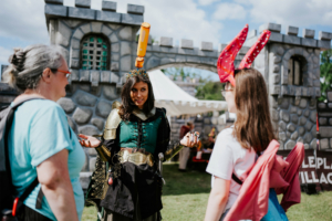 Roleplay Village Ein Kostümierter mit Krone und Rüstung spricht mit zwei Besucherinnen auf dem Elbenwald Festival vor einer mittelalterlichen Kulisse. Die Atmosphäre ist lebhaft, mit bunten Accessoires und einem kreativen Austausch zwischen den Teilnehmern.