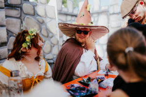 Roleplay Village Bunt kostümierte Teilnehmer am Elbenwald Festival sitzen an einem Tisch, umgeben von kreativen Bastelmaterialien. Ein junger Mann mit einem großen Hut und Sonnenbrille lächelt, während eine Frau mit Blumenkranz neben ihm sitzt.