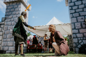 Roleplay Village Zwei Cosplayer auf dem Elbenwald Festival: Ein Charakter in Rüstung mit Schwert konfrontiert einen knienden Charakter in einem braunen Gewand. Im Hintergrund sind Festivalbesucher und Zeltstrukturen sichtbar, die die lebendige Atmosphäre des Events unterstreichen.
