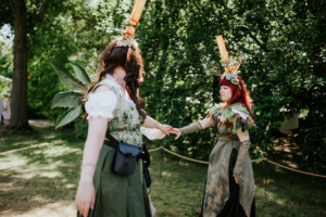 Zwei Frauen in fantasievollen Kostümen mit Pflanzenmotiven und auffälligen Accessoires interagieren auf dem Elbenwald Festival. Die Umgebung ist grün und lebendig, was die Atmosphäre des Festivals unterstreicht.