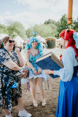 Cosplay-Künstlerinnen beim Elbenwald Festival, lächelnd und im Gespräch. Eine trägt ein farbenfrohes Kostüm mit blauen Haaren und Accessoires, während die andere im klassischen Stil gekleidet ist. Der Hintergrund zeigt Festivalzelte und eine lebhafte Atmosphäre.