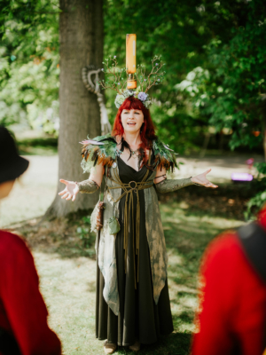 Roleplay Village Eine Frau mit rotem Haar und einem beeindruckenden Kostüm aus Naturmaterialien spricht enthusiastisch auf dem Elbenwald Festival. Ihre Kopfbedeckung aus Ästen und Blumen zieht die Aufmerksamkeit der Festivalbesucher auf sich, während sie eine lebendige Präsentation hält.