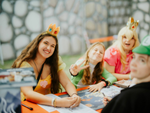 Roleplay Village Vier junge Frauen in bunten Kostümen, darunter Prinzessinnen, lächeln fröhlich an einem Tisch beim Elbenwald Festival. Sie interagieren spielerisch, während sie an einem Brettspiel arbeiten, umgeben von einer festlichen Atmosphäre.