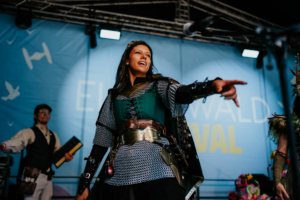Eine PerformerIn in Rüstung steht auf der Bühne des Elbenwald Festivals, während sie das Publikum mit einem enthusiastischen Fingerzeig anspricht. Im Hintergrund sind weitere Künstler und eine farbenfrohe Dekoration zu sehen.