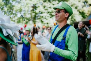 Ein Mann in einem grünen Luigi-Kostüm mit weißen Handschuhen applaudiert bei einem Auftritt auf dem Elbenwald Festival. Im Hintergrund sind weitere kostümierte Festivalbesucher zu sehen, die die festliche Atmosphäre genießen.