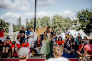 Percy Jackson Talk Eine Moderatorin spricht auf der Bühne beim Elbenwald Festival, umgeben von einem begeisterten Publikum in Kostümen. Die Atmosphäre ist lebhaft und kreativ, mit Teilnehmern, die verschiedene Charaktere aus der Fantasy-Welt darstellen.