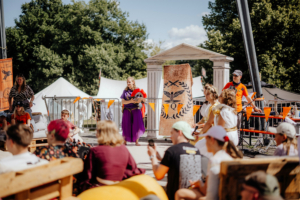 Percy Jackson Talk Bühne beim Elbenwald Festival mit kostümierten Darstellern, darunter eine Frau in lila Gewand, die ein Musikinstrument hält. Zuschauer sitzen auf Holzpaletten und genießen die Aufführung im Freien, umgeben von bunten Fahnen und Zelten.