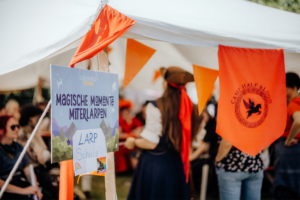 Bunte Fahnen und ein Schild mit der Aufschrift „Magische Momente mit LARP“ kennzeichnen einen Bereich des Elbenwald Festivals, wo Teilnehmer in fantasievolle Rollenspiele eintauchen können. Im Hintergrund sind begeisterte Besucher zu sehen.