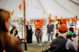 Percy Jackson Talk Vier Frauen stehen in einem Zelt beim Elbenwald Festival, umgeben von orangefarbenen Fähnchen. Sie interagieren mit dem Publikum, das in der Nähe sitzt und interessiert zuschaut. Die Atmosphäre ist lebhaft und einladend.