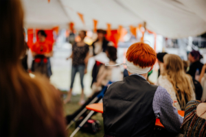Ein Teilnehmer des Elbenwald Festivals mit rotem Haar, der in einem Zelt sitzt und auf eine Darbietung blickt. Im Hintergrund sind weitere Festivalbesucher zu sehen, die sich an Aktivitäten beteiligen, umgeben von bunten Dekorationen.