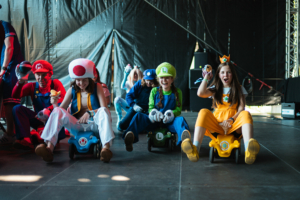 Mario Kart Turnier-Finale Spaßige Szene auf dem Elbenwald Festival: Teilnehmer in bunten Kostümen, darunter Mario, Luigi und Prinzessin Peach, fahren in kleinen Karts und zeigen ihre Freude an der Veranstaltung. Die Atmosphäre ist lebhaft und voller Nostalgie für Videospiel-Fans.