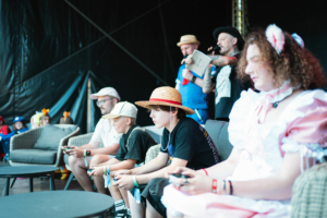 Mario Kart Turnier-Finale Teilnehmer des Elbenwald Festivals sitzen auf einer Couch und spielen Videospiele. Im Hintergrund sind Zuschauer und Moderatoren zu sehen. Die Atmosphäre ist lebhaft und zeigt die Verbindung von Gaming und Gemeinschaft auf dem Festival.